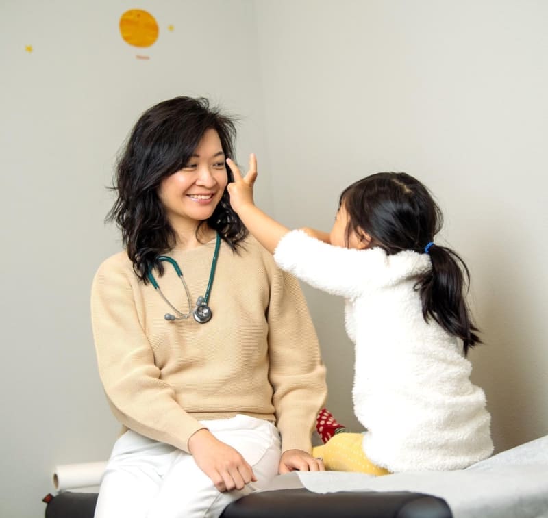 dr-chung-with-young-girl-patient-in-exam-room-happy-bun-peds. Dr. Jennie Chung smiling warmly as a young girl in a white fuzzy jacket touches her face playfully on a pediatric exam table.