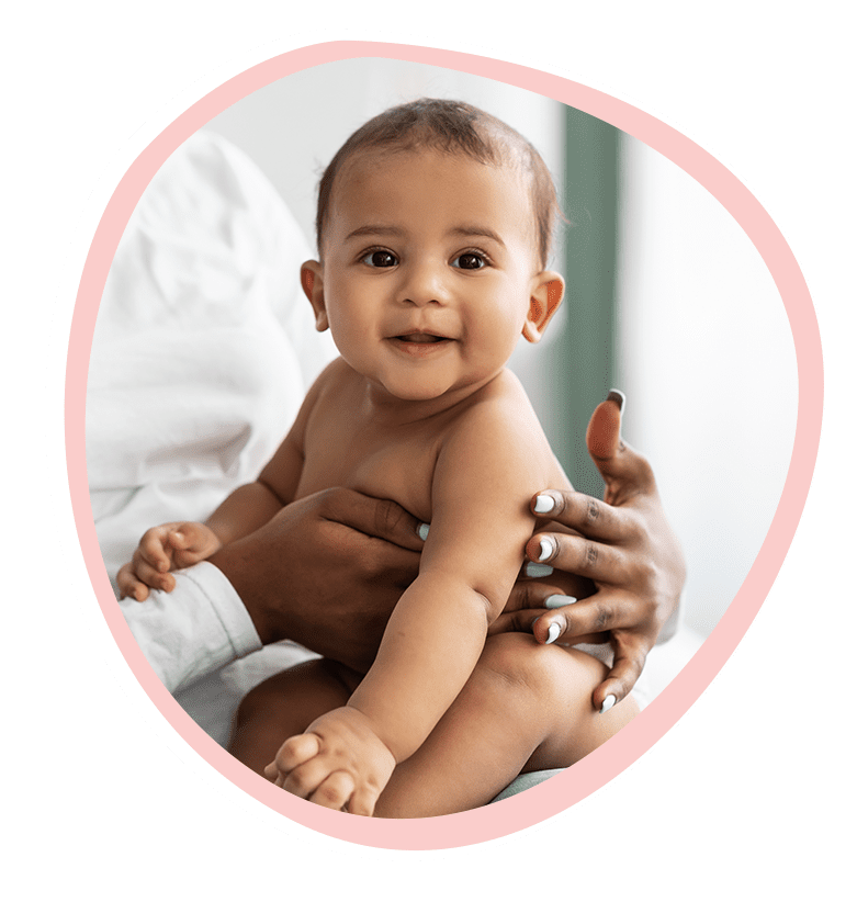 smiling-baby-held-upright-in-pink-circle-frame-happy-bun-peds. A smiling, bright-eyed baby being held upright by a caregiver's hands, displayed in a soft pink organic circle frame on a black background.