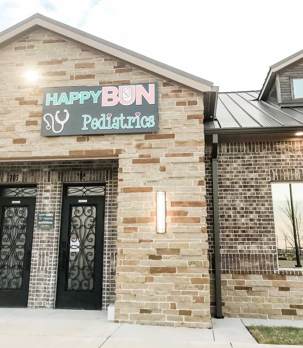 happy-bun-pediatrics-office-building-exterior-happy-bun-peds. The exterior of the Happy Bun Pediatrics office building, a brick commercial building with a dark sign displaying the practice name and logo.