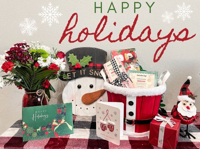 happy-holidays-festive-display-happy-bun-peds. A festive holiday table display with a snowman sign, flower bouquet, Santa bucket, holiday cards, and a small Santa figurine, with "Happy Holidays" text above.