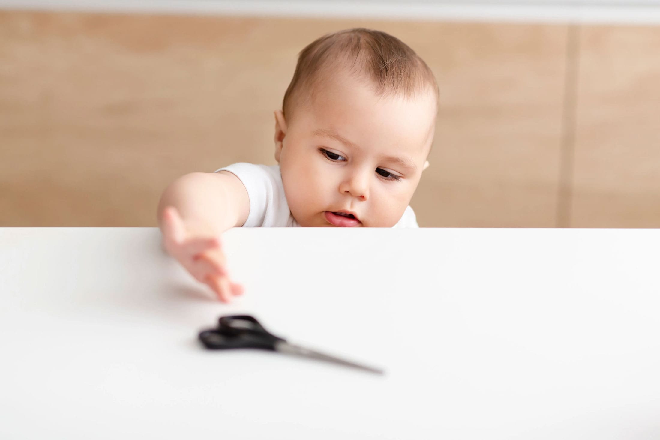 baby-reaching-for-scissors-safety-hazard-happy-bun-peds. A curious baby in a white onesie reaching over the edge of a white table toward a pair of scissors, illustrating a common household safety hazard.