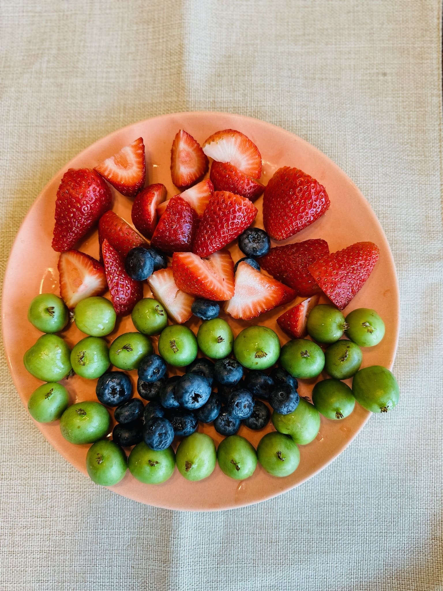 overhead-plate-of-fresh-strawberries-blueberries-and-green-grapes-happy-bun-peds. An overhead view of a pink plate filled with sliced strawberries, blueberries, and small green grapes on a linen surface.
