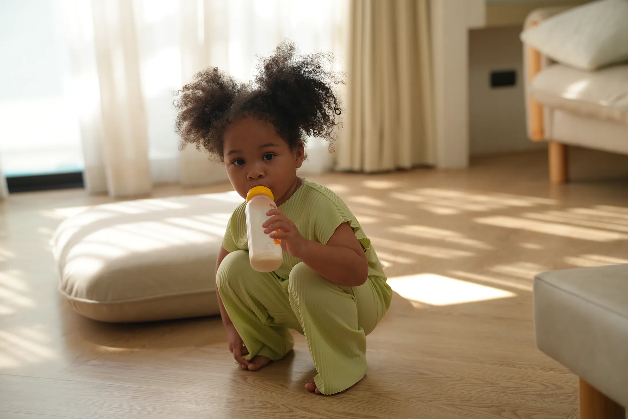 toddler-drinking-from-bottle-on-sunlit-floor-happy-bun-peds. Toddler with curly pigtails squatting on a wooden floor while drinking from a baby bottle in a sunlit room.