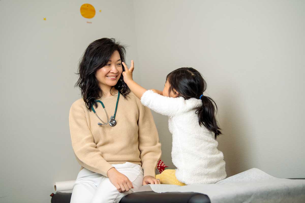 dr-chung-with-young-girl-patient-in-exam-room-happy-bun-peds. Dr. Jennie Chung smiling warmly as a young girl in a white fuzzy jacket touches her face playfully on a pediatric exam table.